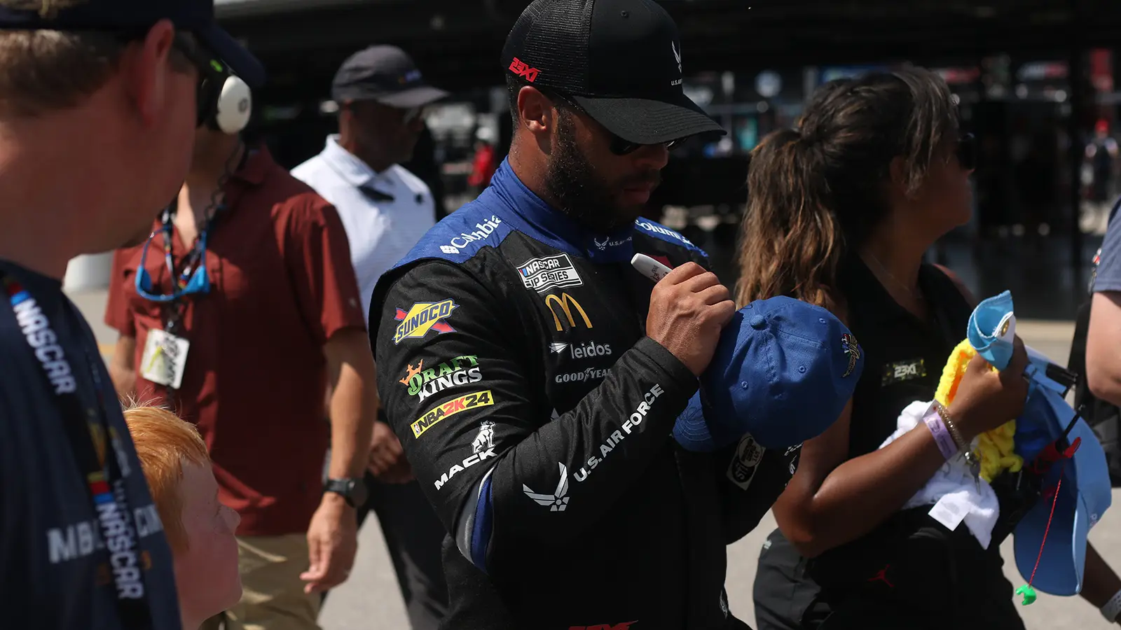Bubba Wallace stands in his fire suit holding a sharpie. He signs the back of a fan's shirt.