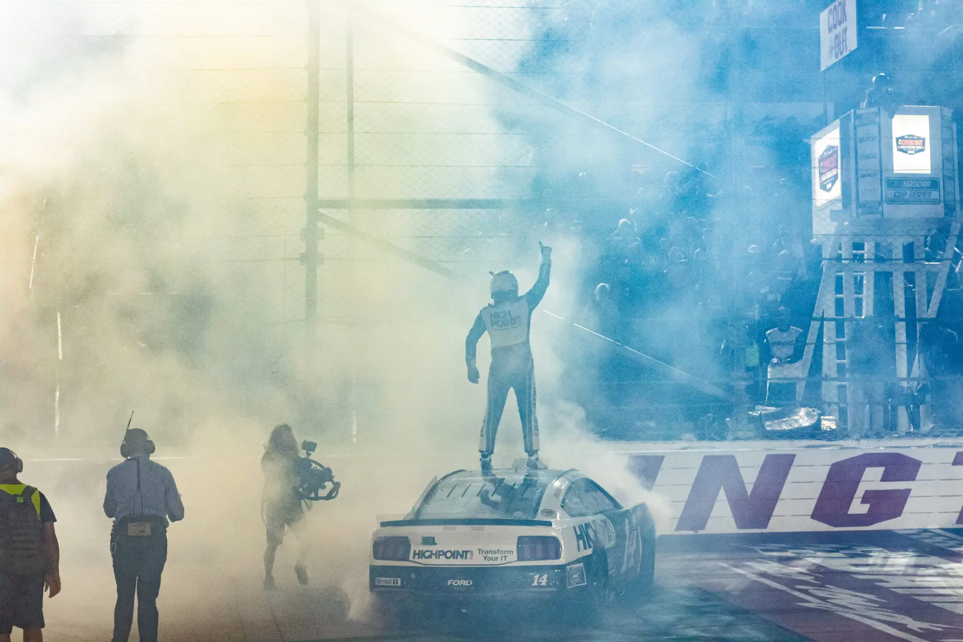 In a smoky image, Chase Briscoe stands on top of his No. 14 black and white race car facing a cheering crowd of fans after his thrilling Darlington Raceway win.
