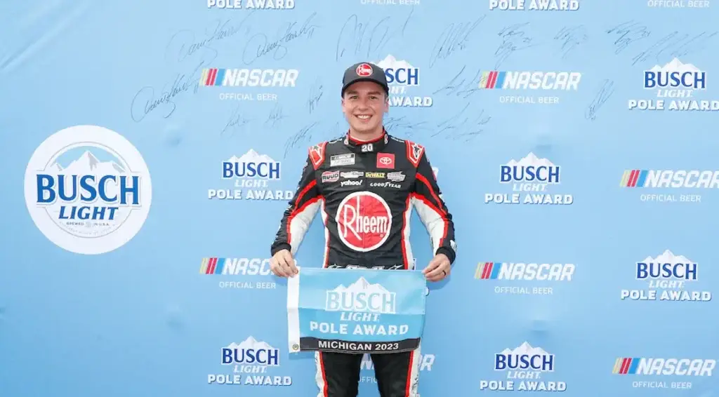 Christopher Bell stands against a light blue background with the Busch Beer and NASCAR logos for the Busch Light Pole Aware. He also holds a light blue flag that says "Busch Light Pole" on it for the race at Michigan International Speedway.