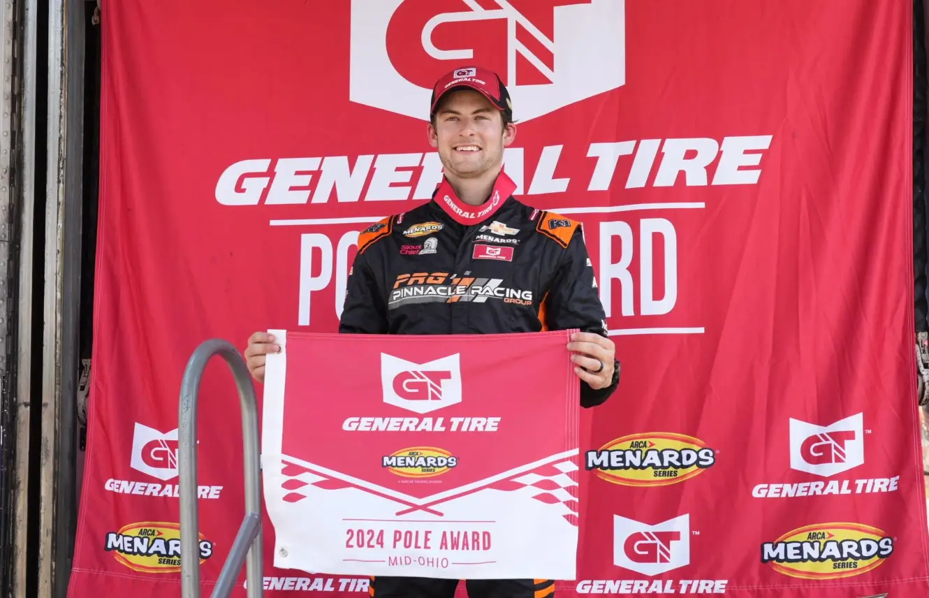 Connor Mosack standing in front of a red General Tire background holding a General Tire Pole Award flag at Mid-Ohio.