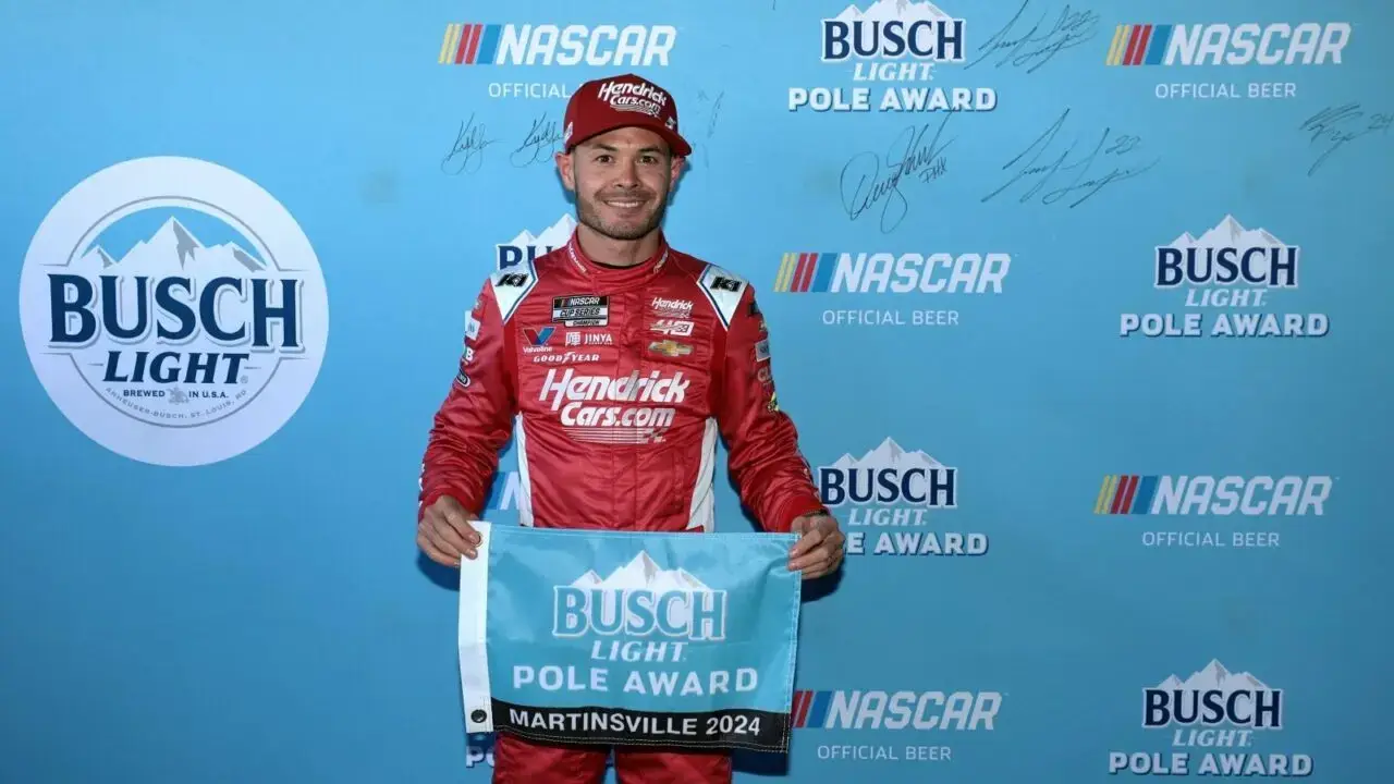 Kyle Larson in his ruby red HendrickCars.com fire suit to celebrate Hendrick Motorsport's 50th anniversary. He's holding a Busch Light Pole Award flag in front of a light blue backdrop.