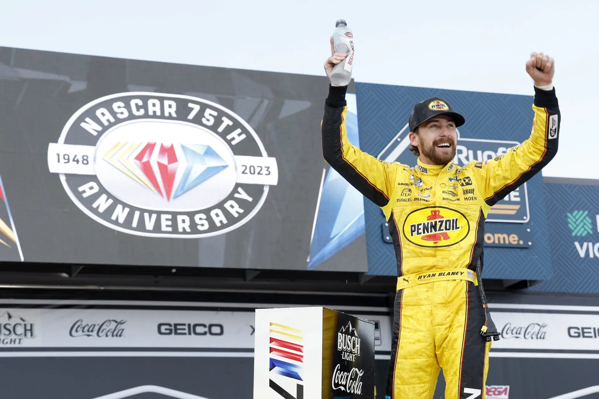 Ryan Blaney in a bright yellow and black Pennzoil fire suit celebrating in victory lane.