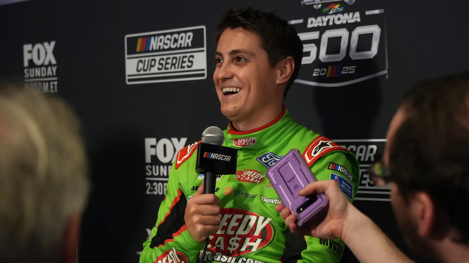 Zane Smith sits smiling in a green and red Speedy Cash fire suit at Daytona 500 media day.