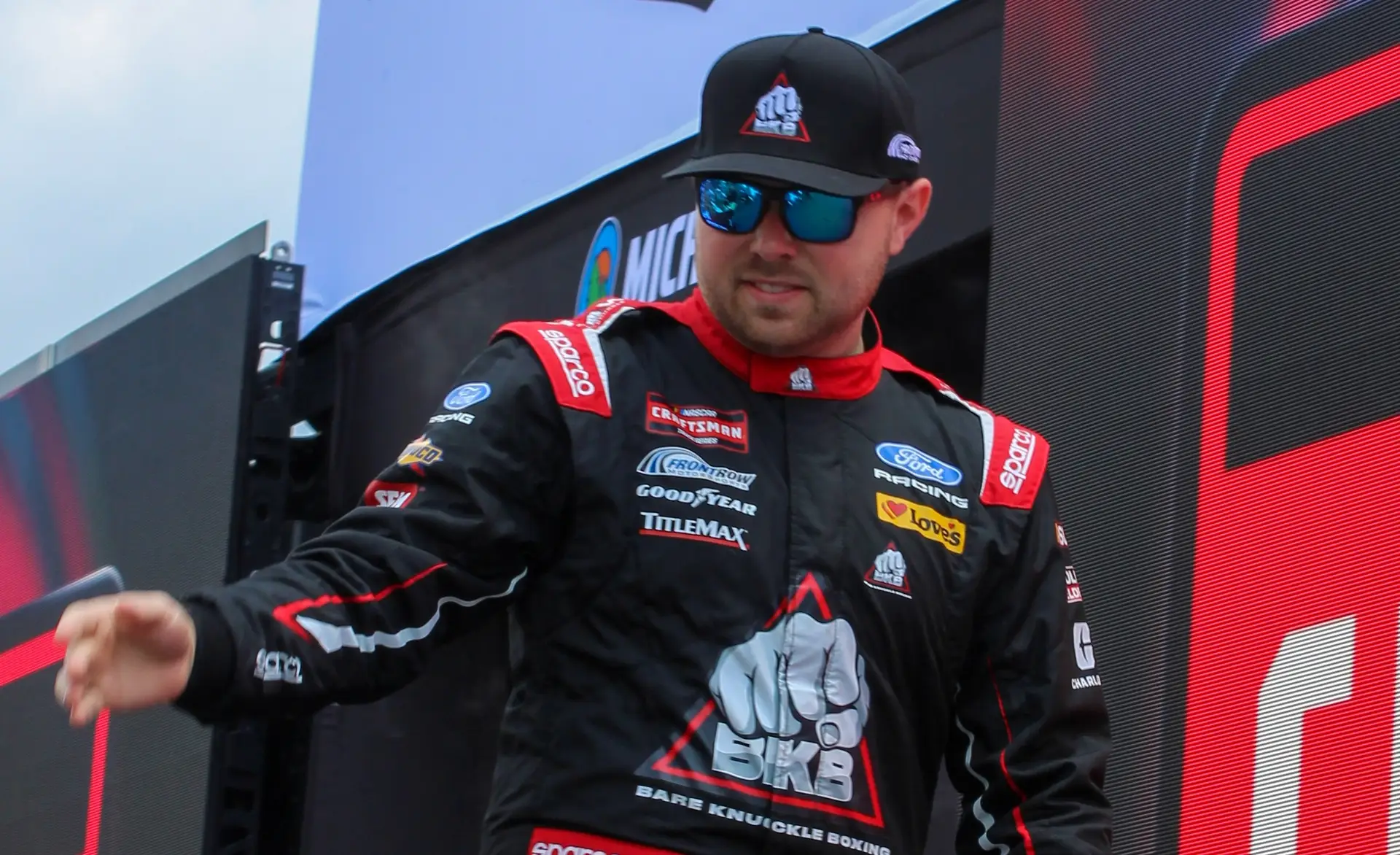 Layne Riggs walks on the driver intro stage at Michigan International Raceway surrounded by screens with the NASCAR Craftsman Truck Series logo on them. He wears a black and red firesuit with black sunglasses and baseball cap.